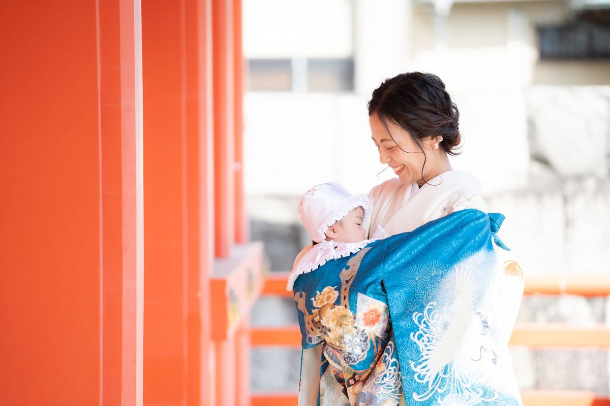 神社の朱色の柱の前で晴れ着の赤ちゃんを抱くママ